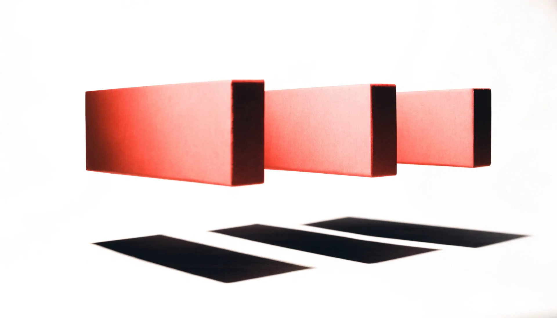Conceptual still life: three coral blocks held in a row above an off-white sweep, each casting its own hard shadow, reading as one form.